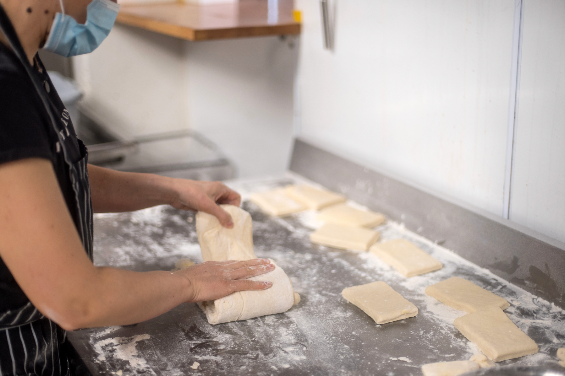 A picture showing a masked person making dough and cutting them into square pieces.