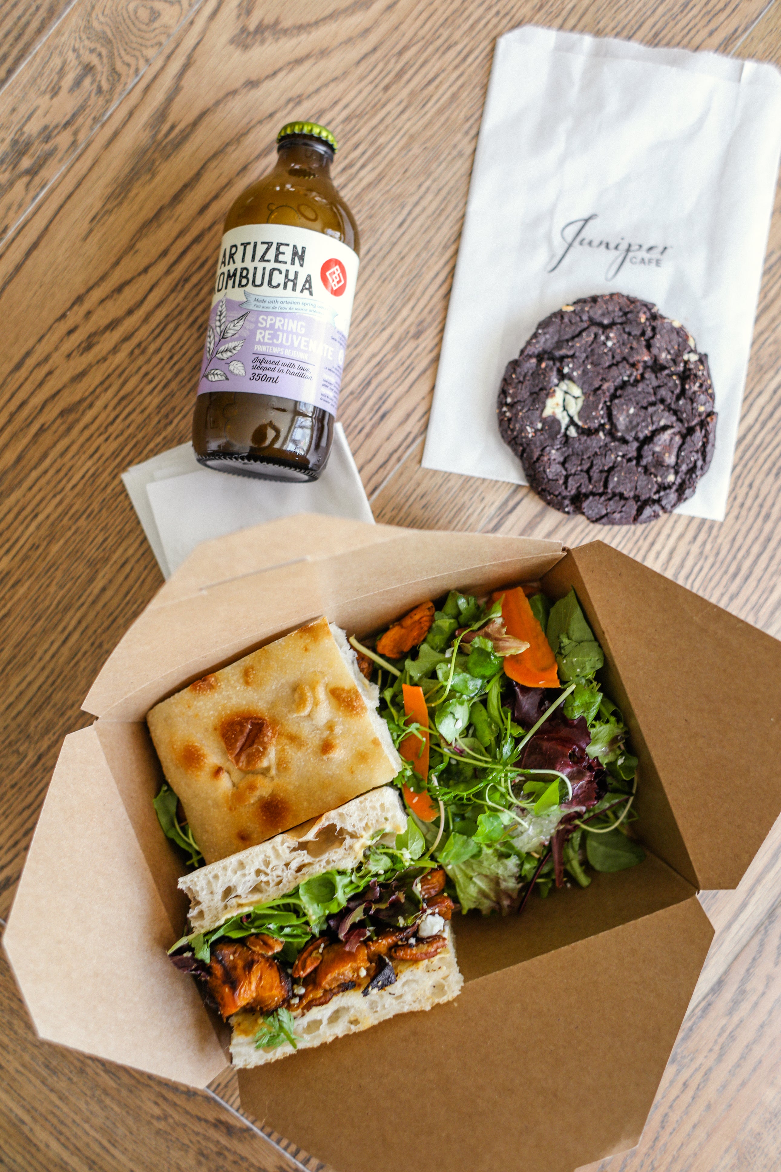 An image showing Kombucha Drink, A Cookie, and a boxed lunch with two sandwiches and a salad served by Juniper Cafe.